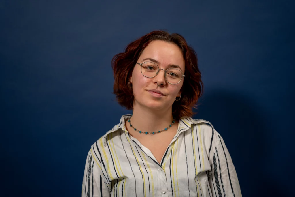 En person med kort, bølget brunt hår, briller og en stribet skjorte står foran en ensfarvet mørkeblå baggrund og ser mod kameraet med et let smil.