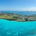 Luftfoto af en frodig, grøn ø omgivet af turkisfarvet vand og koralrev, med huse og bygninger spredt ud over landskabet under en klar blå himmel.