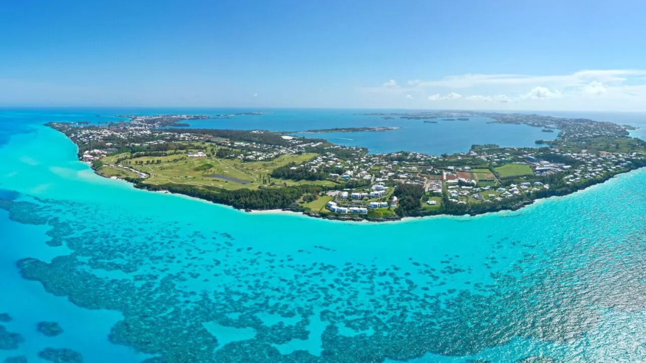 Luftfoto af en frodig, grøn ø omgivet af turkisfarvet vand og koralrev, med huse og bygninger spredt ud over landskabet under en klar blå himmel.