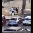 En mand og en kvinde har et skænderi på et fortov nær parkerede biler og skraldespande, med et picnicbord og græs i baggrunden.