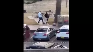 En mand og en kvinde har et skænderi på et fortov nær parkerede biler og skraldespande, med et picnicbord og græs i baggrunden.