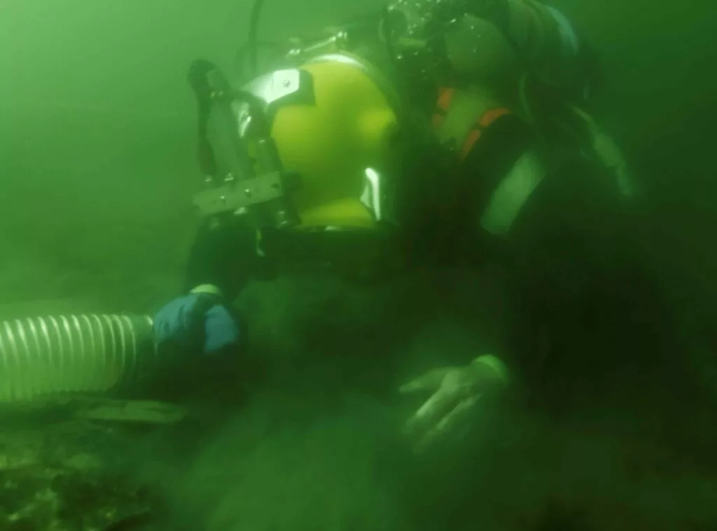 En undervandsdykker iført gul hjelm og dragt bruger en stor vakuumslange til at fjerne sediment fra havbunden, hvilket skaber en sky af affald i det grøntonede vand.