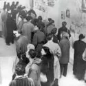 Sort-hvidt foto af et overfyldt kunstgalleri, hvor folk i vinterfrakker ser på fotos og kunstværker, der er hængt op på væggene. Scenen antyder en udstilling eller et offentligt arrangement med deltagere i forskellige aldre.