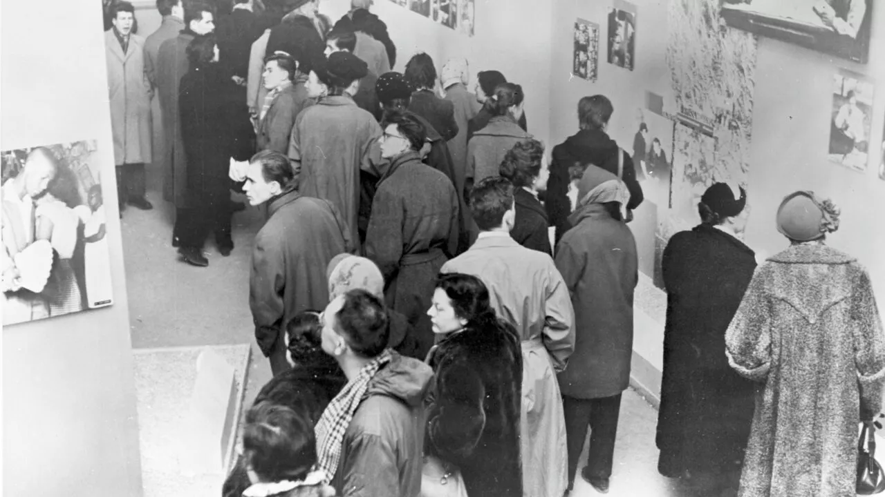 Sort-hvidt foto af et overfyldt kunstgalleri, hvor folk i vinterfrakker ser på fotos og kunstværker, der er hængt op på væggene. Scenen antyder en udstilling eller et offentligt arrangement med deltagere i forskellige aldre.