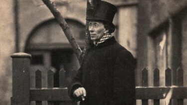 En mand i mørk frakke og høj hat, der ligner H.C. Andersen, står ved siden af et træhegn og holder en stok med en sløret bygning og et træ i den sepiatonede baggrund. Billedet virker gammelt og tidløst.