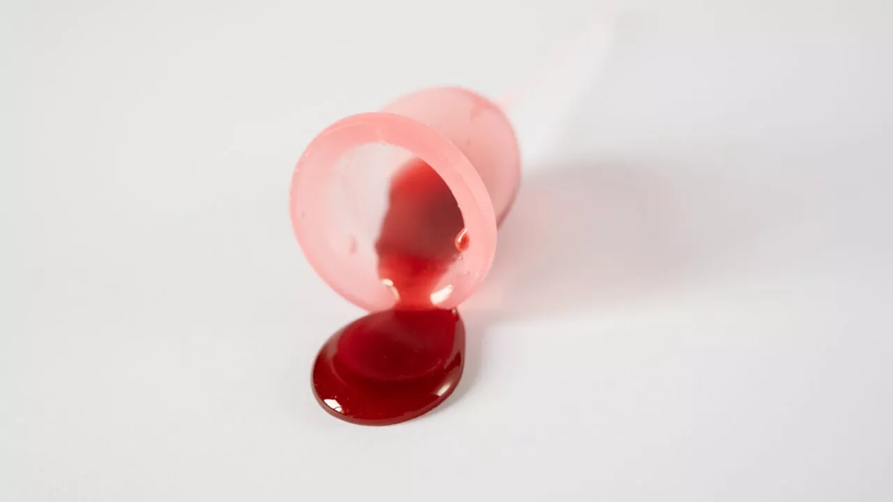 En lyserød menstruationskop, der er væltet på en hvid overflade, med mørkerød væske, der løber ud af den.