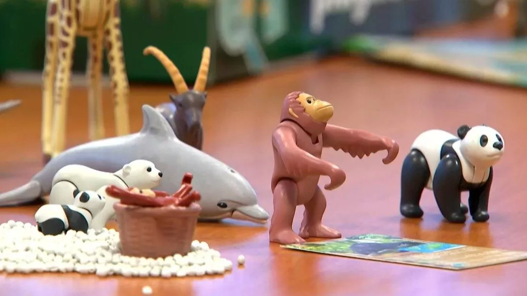 Legetøjsdyrfigurer, herunder en delfin, pandabjørne, en abe, en giraf og en antilope, er anbragt på en bordplade med en kurv, nogle hvide perler og et spillekort i nærheden.