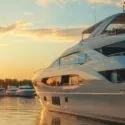 En stor, moderne hvid yacht ligger i en marina ved solnedgang med roligt vand, der reflekterer den gyldne himmel, og flere andre både er synlige i baggrunden.