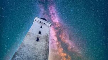 Et højt stenfyrtårn står på en nattehimmel fyldt med stjerner og Mælkevejens livlige, farverige striber. Billedet er taget fra en lav vinkel, hvilket understreger fyrtårnets højde.