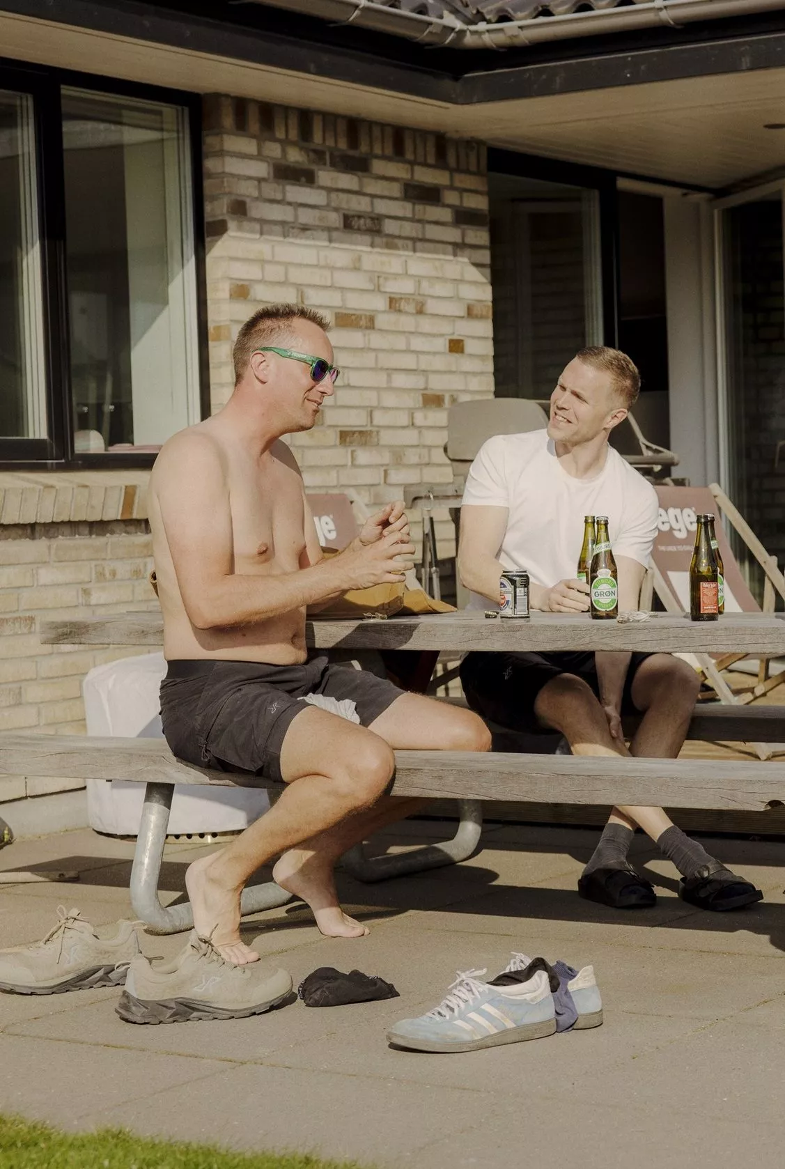 To mænd sidder på en picnicbænk uden for et murstenshus og slapper af med drinks. Den ene mand har bar overkrop og solbriller på, mens den anden har en hvid T-shirt på. Sko og sokker ligger spredt på jorden i nærheden.