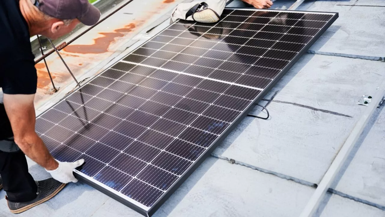 To arbejdere i handsker og arbejdstøj installerer et stort solpanel på et tag og justerer det omhyggeligt. I baggrunden ses en del af taget og sikkerhedsudstyr.