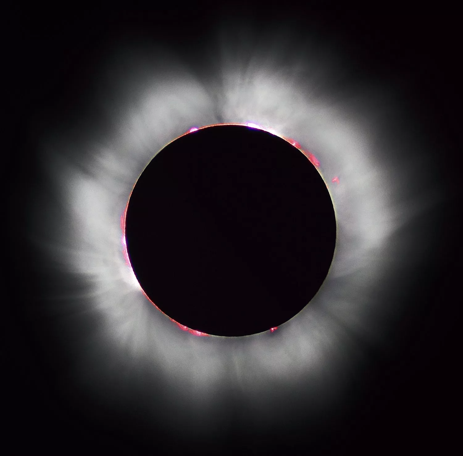 En total solformørkelse, hvor månen fuldstændigt dækker solen, omgivet af solens glødende hvide korona og røde soludbrud, der kan ses langs kanten.
