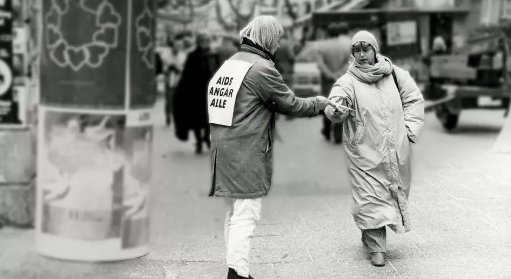 En person iført en frakke med et skilt med teksten AIDS ANGÅR ALLE rækker en avis til en anden person klædt i lang frakke og tørklæde på en travl gade i byen. Baggrunden er sløret.