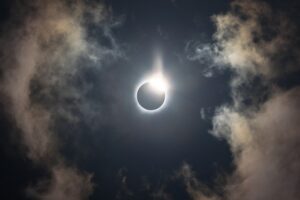 En solformørkelse med solen delvist dækket af månen, hvilket skaber en lysende korona og diamantringseffekt, omgivet af en mørk himmel og tynde, oplyste skyer.