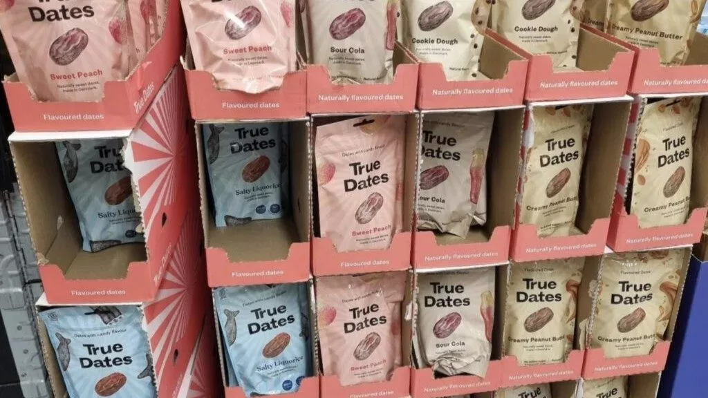 Flere poser med True Dates-dadler med smag står på hylderne. Smagsvarianterne omfatter Sweet Peach, Sour Cola, Salty Licorice, Cookie Dough og Crispy Peanut Butter. Emballagen er i pastelfarver.