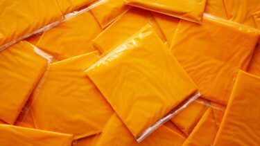 Individuelle skiver af forarbejdet orange ost er pakket ind i klar plast og arrangeret i et overlappende mønster, der fylder hele billedet.