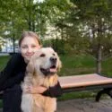 En ung person med lyst hår og en sort top sidder på en bænk i det fri og krammer en glad golden retriever. Grønne træer og græs omgiver dem og skaber et fredeligt parkområde.