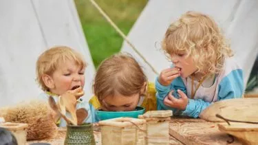 Tre små børn sidder ved et rustikt træbord udendørs og spiser snacks. Et barn kigger ned i en skål, mens de andre bruger deres hænder til at spise med. I baggrunden ses campingtelte og grønt græs.