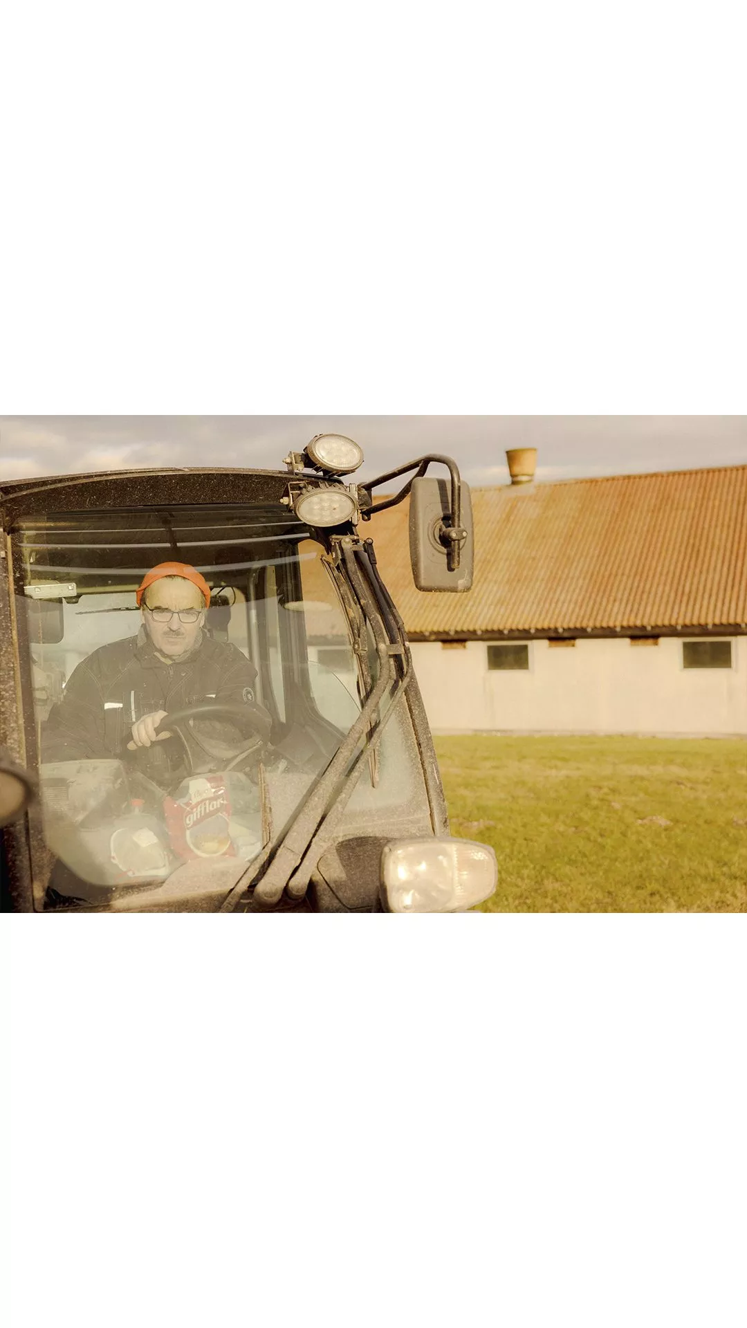 En person med briller og orange pandebånd sidder i førersædet på en beskidt traktor, der er parkeret på græs foran en bygning med rødt tegltag.