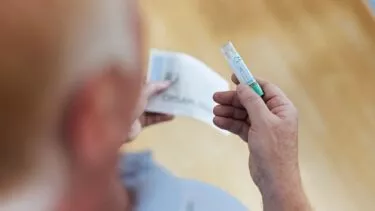 En person holder et lille plastikglas med en grøn hætte i den ene hånd og et foldet instruktionsark i den anden, set ovenfra. Baggrunden er ude af fokus.