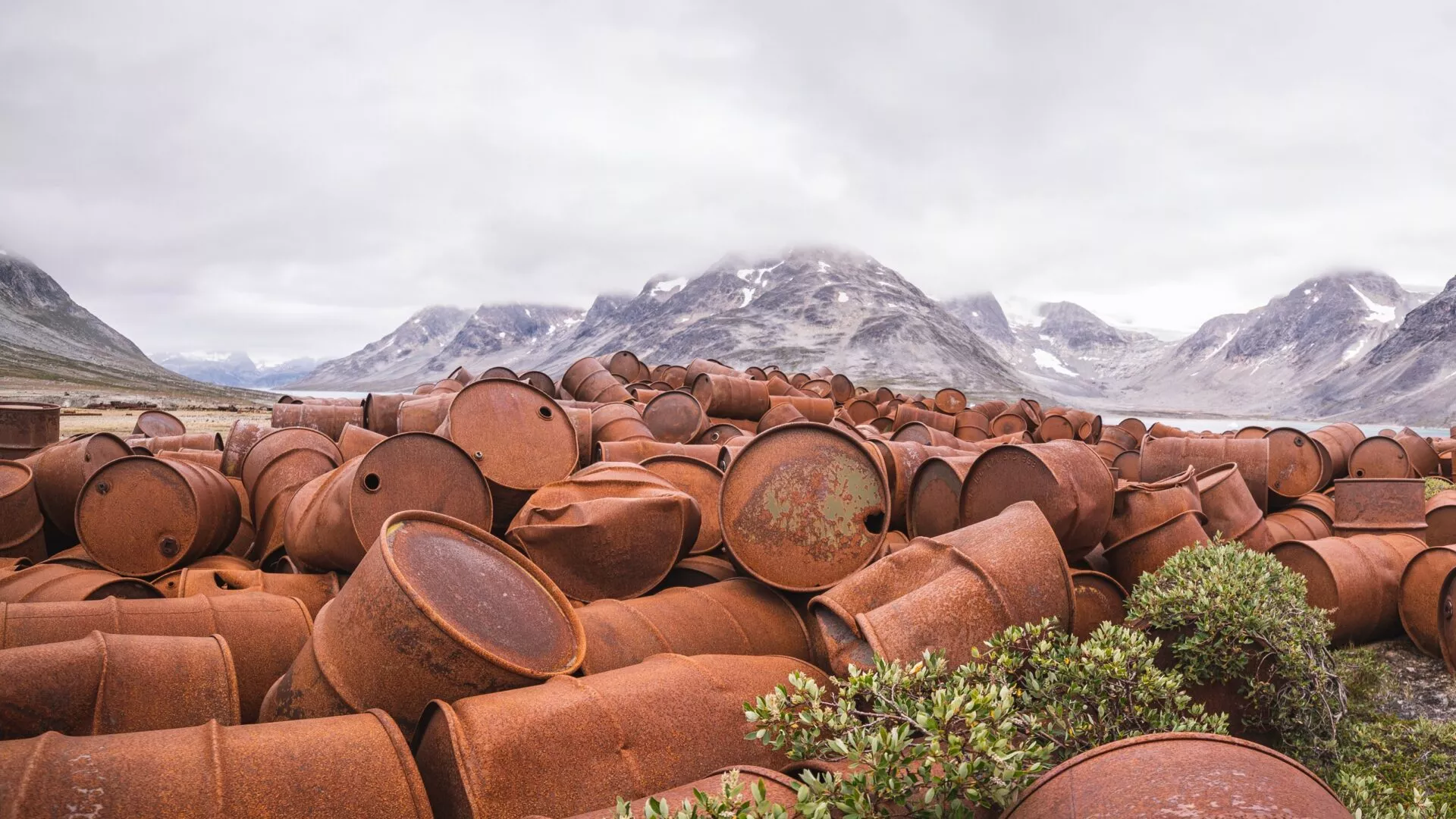 En stor bunke rustne metaltønder stablet udendørs i et goldt landskab med bjerge og overskyet himmel i baggrunden; nogle grønne buske er synlige i forgrunden.