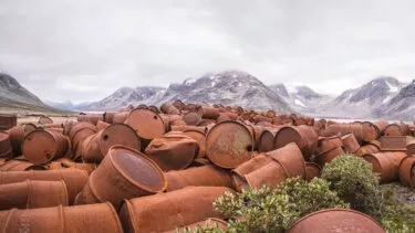 En stor bunke rustne metaltønder stablet udendørs i et goldt landskab med bjerge og overskyet himmel i baggrunden; nogle grønne buske er synlige i forgrunden.