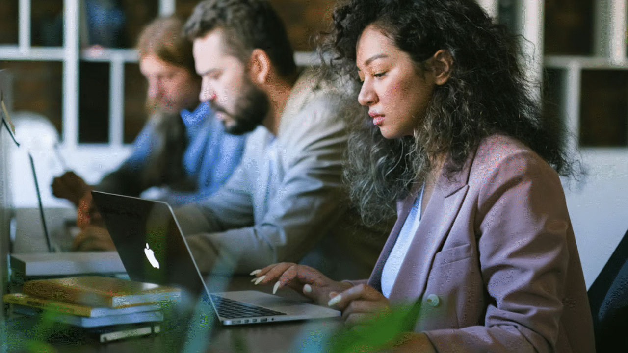 Tre personer sidder ved et skrivebord og arbejder på bærbare computere på et kontor. Fokus er på en kvinde med krøllet hår og lyserød blazer, som skriver, mens to andre personer arbejder i baggrunden. Bøger og grønne planter er også synlige.