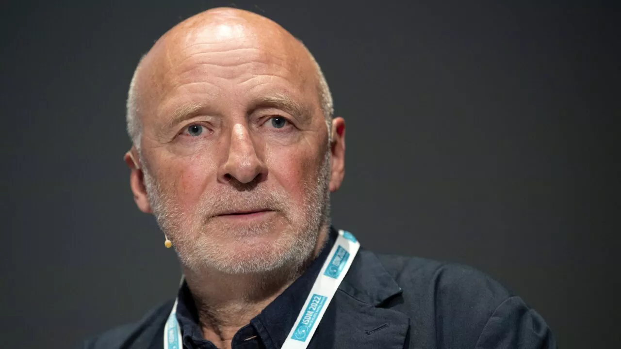 En ældre mand med skaldet hoved og gråt skæg iført en mørk jakke og en hvid snor taler med et alvorligt udtryk mod en almindelig mørk baggrund.