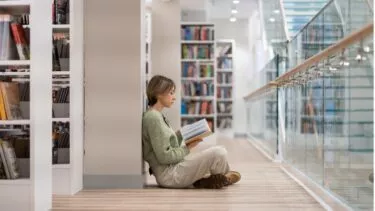 En person sidder på gulvet i et bibliotek, læner sig op ad en væg og læser en bog. Bogreoler og et glasgelænder omkranser det lyse, moderne rum.