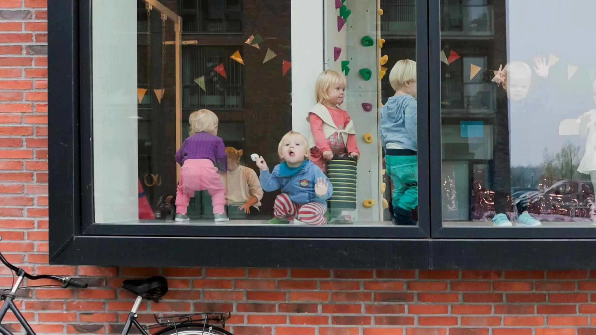 Flere små børn står og leger bag et stort vindue og kigger ud. Rummet er dekoreret med farverige flag, og en cykel er parkeret op ad den røde murstensvæg under vinduet.