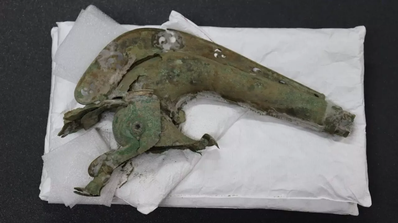 En korroderet, antik pistol med grønlig patina ligger på et hvidt, polstret klæde mod en sort baggrund. Pistolen ser gammel og forvitret ud med rust og manglende dele.