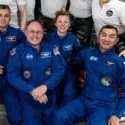 Fire astronauter i blå flyverdragter sidder og knæler sammen og smiler til et gruppebillede inde i et rumfartøj. Yderligere tre personer i hvide skjorter står bag dem, delvist synlige. Rumudstyr ses i baggrunden.