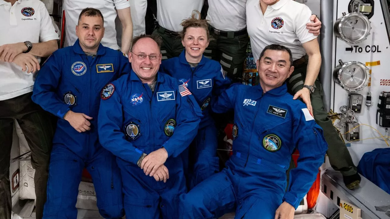 Fire astronauter i blå flyverdragter sidder og knæler sammen og smiler til et gruppebillede inde i et rumfartøj. Yderligere tre personer i hvide skjorter står bag dem, delvist synlige. Rumudstyr ses i baggrunden.