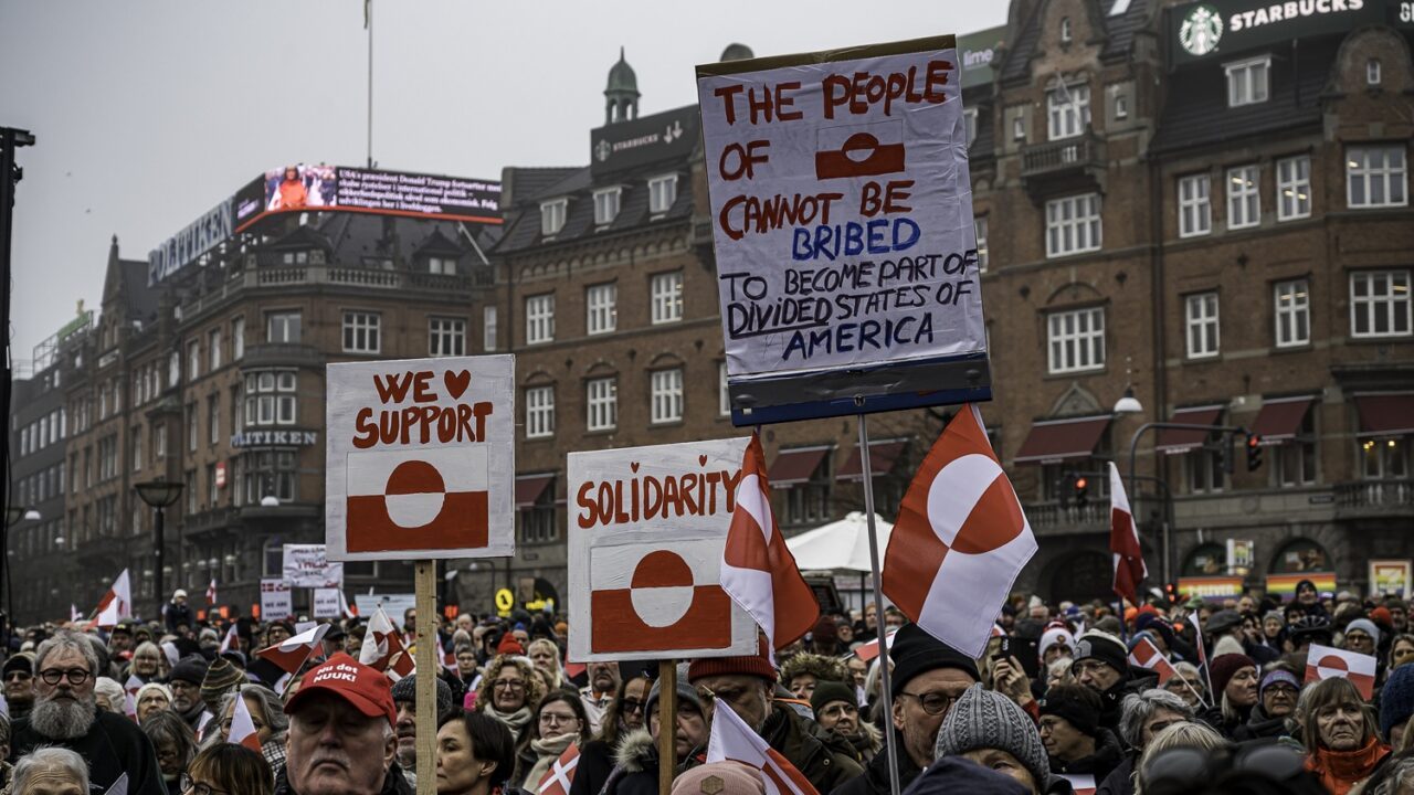 En stor menneskemængde holder grønlandske flag og protestskilte med budskaber som "Solidaritet", "Vi støtter" og "Det grønlandske folk kan ikke bestikkes til at blive en del af de splittede stater i Amerika" på et torv i byen.