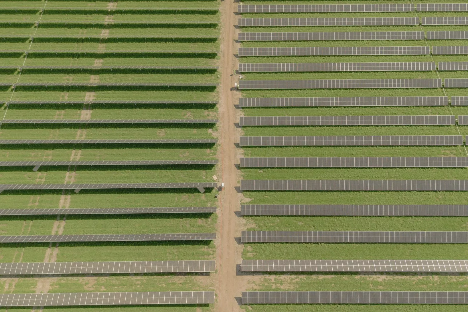 Luftfoto af en solcellepark med rækker af solcellepaneler jævnt fordelt på grønt græs, adskilt af jordstier, der løber lodret og vandret gennem marken.