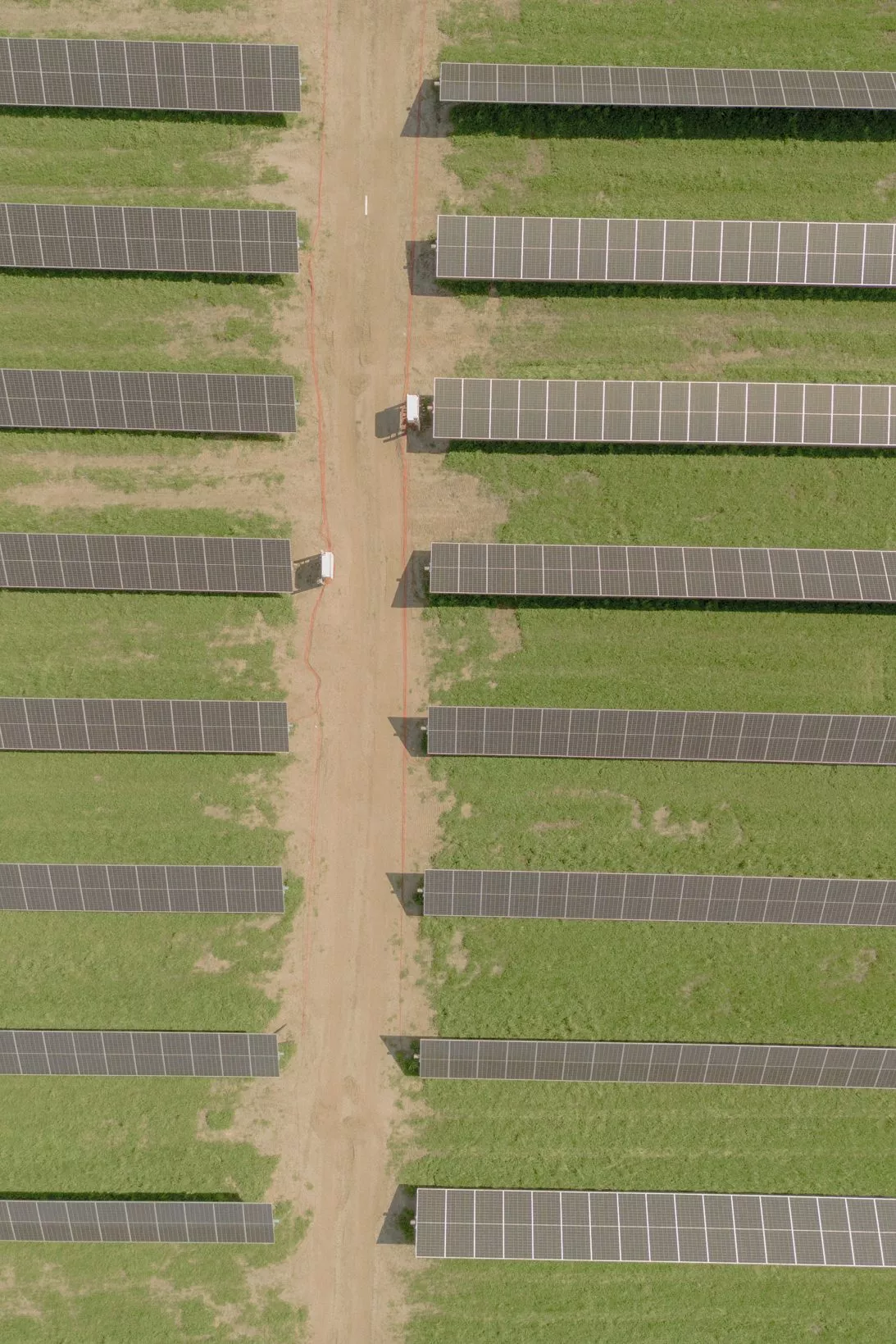 Luftfoto af rækker af solcellepaneler på grønt græs, adskilt af jordstier, på en solcellepark. To hvide køretøjer er parkeret på den centrale sti mellem panelerne.