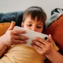 En ung dreng i en gul skjorte sidder i en sofa og holder en hvid smartphone med begge hænder, mens han fokuserer på skærmen. Orange og mørke puder er synlige bag ham.