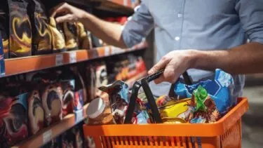 En person i blå skjorte handler i en dagligvarebutik med en orange kurv fyldt med forskellige emballerede fødevarer og rækker ud efter et produkt på en hylde.