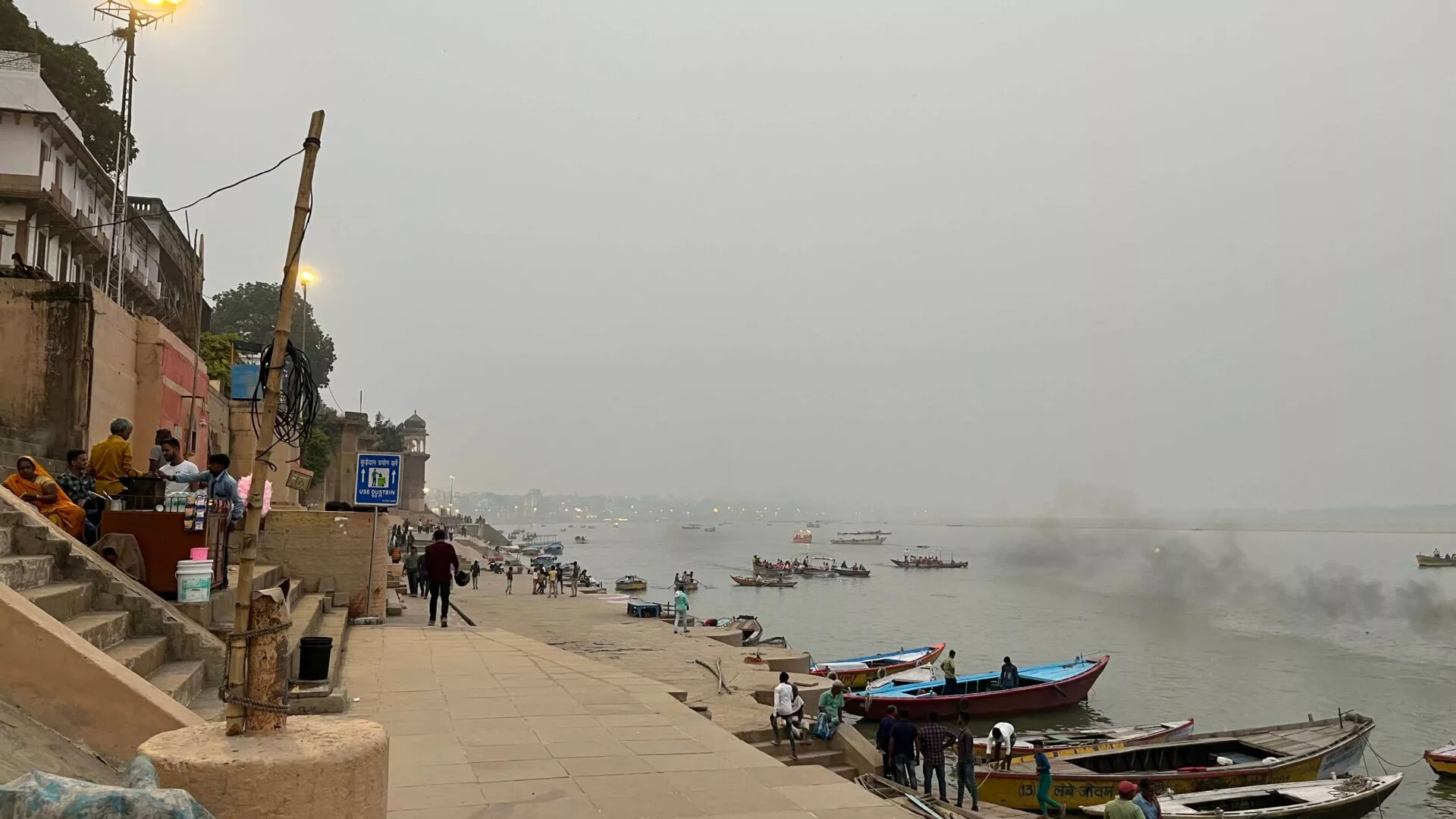 Folk sidder og går langs en ghat med stentrapper ved floden i Varanasi, Indien. Både ligger for anker ved flodbredden, og røg stiger op i det fjerne under en diset himmel. Nogle lanterner og bygninger står langs venstre side.