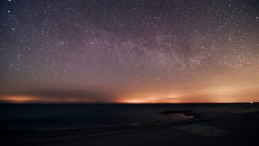 En stjernefyldt nattehimmel strækker sig over et roligt, mørkt hav med et svagt orange skær i horisonten og et blødt lys, der reflekteres i vandet og på kysten.