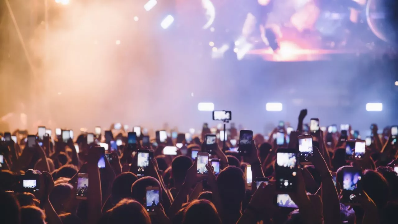 Et stort publikum til en koncert holder smartphones op for at optage og tage billeder, med en stærkt oplyst scene og spotlights synlige gennem røg i baggrunden.