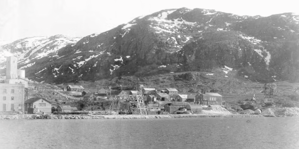 Sort-hvidt foto af en lille landsby med spredte bygninger og strukturer ved foden af et klippefyldt, sneklædt bjerg ved siden af et vandløb.