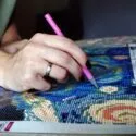 En person bruger en pink stylus til at placere små farvede perler på et mosaiklærred, der ligner Van Goghs Starry Night-mønster i et diamantmaleri.