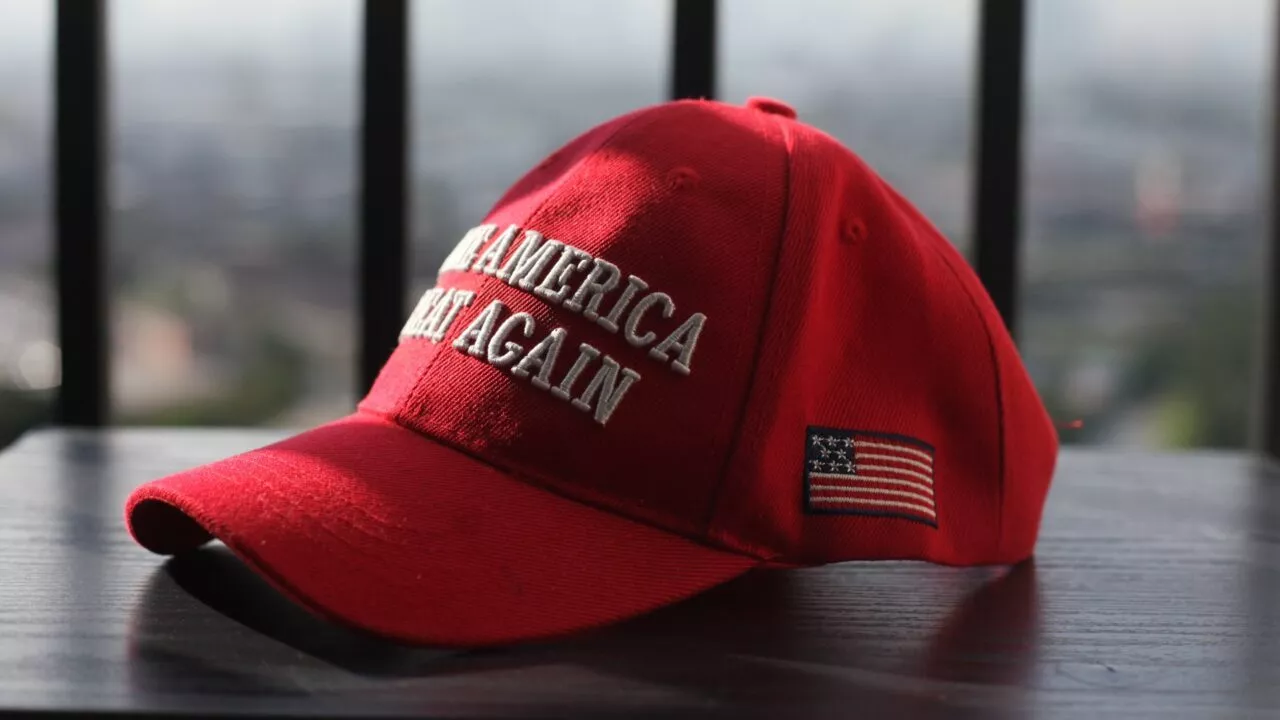 En rød baseballkasket med hvide broderede bogstaver, hvor der står MAKE AMERICA GREAT AGAIN, og et lille amerikansk flagmærke på siden, hviler på en mørk overflade med slørede lodrette streger i baggrunden.