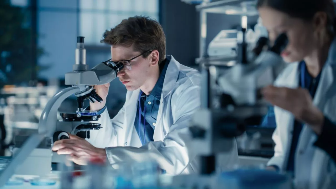 To forskere i hvide kitler bruger mikroskoper i et laboratorium. Den ene mand observerer nøje en prøve, mens en kvinde arbejder i baggrunden. Omgivelserne er fyldt med laboratorieudstyr og har en fokuseret, professionel atmosfære.