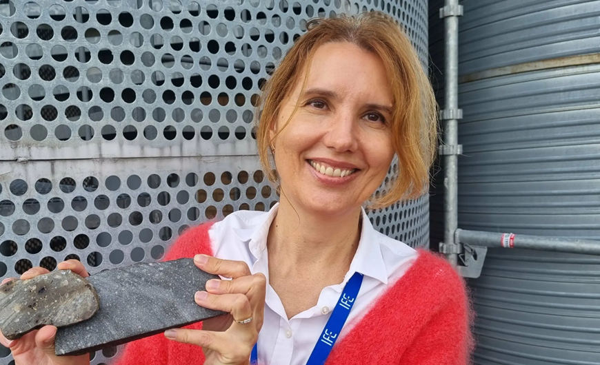 En smilende kvinde med lysebrunt hår holder to sten i hænderne. Hun er iført en rød sweater, en hvid skjorte med krave og en blå snor. En metallisk, perforeret baggrund er synlig bag hende.