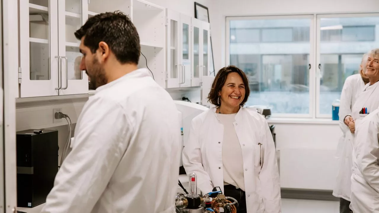 Tre personer i hvide kitler arbejder og interagerer i et lyst laboratorium. En kvinde i midten smiler, mens en anden person er i baggrunden, og en mand er fokuseret på udstyr i forgrunden.