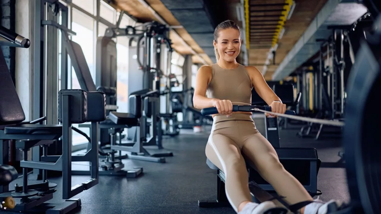 En kvinde i beige træningstøj smiler, mens hun bruger en romaskine i et rummeligt, moderne fitnesscenter med forskelligt træningsudstyr i baggrunden.