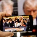 En hånd holder en smartphone, der viser en direkte udsendelse af Donald Trumps præsidentindsættelse, med Trumps forstørrede billede sløret på en skærm i baggrunden.