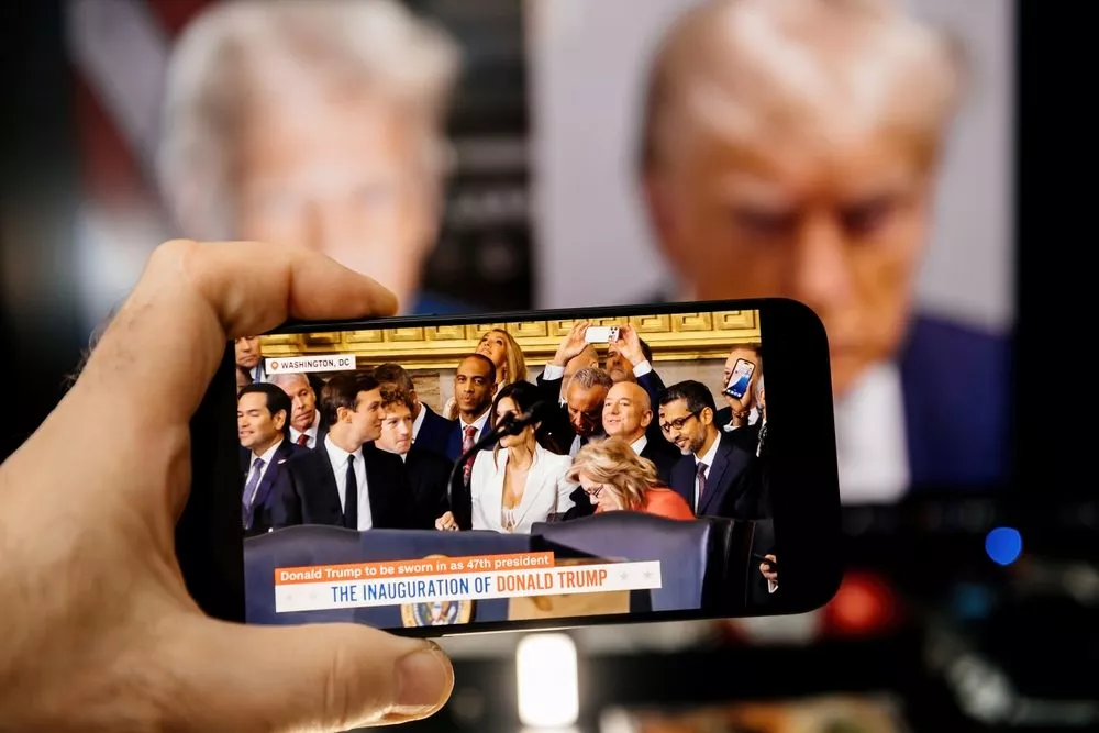 En hånd holder en smartphone, der viser en direkte udsendelse af Donald Trumps præsidentindsættelse, med Trumps forstørrede billede sløret på en skærm i baggrunden.